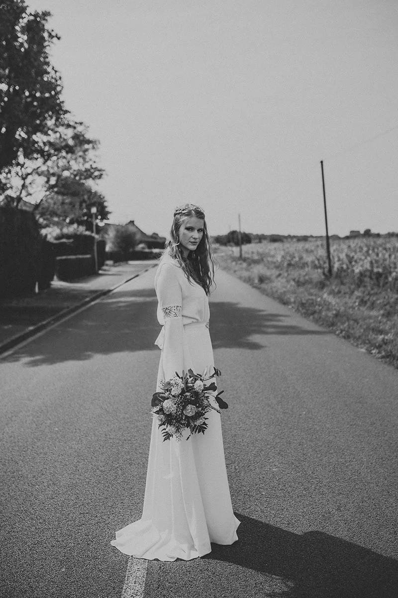 Mariée en robe blanche bohème avec bouquet, posant seule sur une route de campagne en noir et blanc.