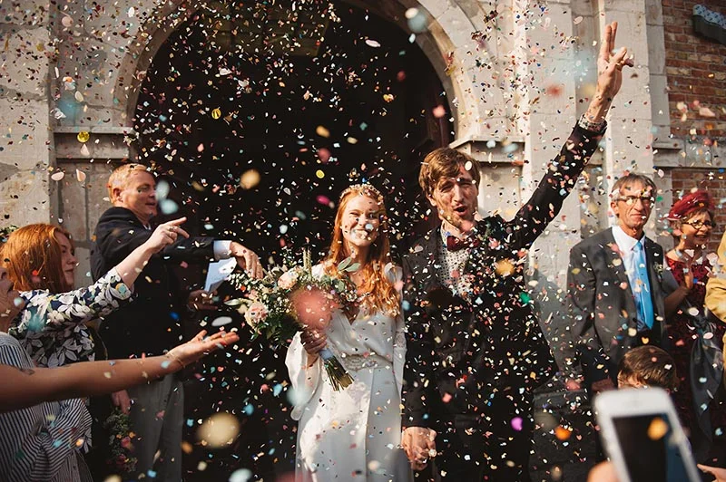Jeunes mariés souriants sous une pluie de confettis colorés à la sortie de l'église, entourés de leurs invités.