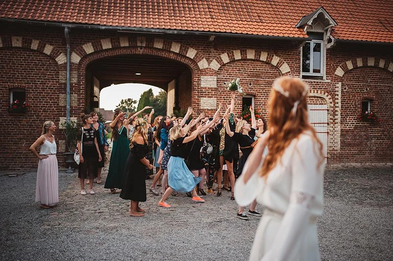 Des invitées lancent un bouquet à une mariée devant une grange en briques dans une ambiance festive et champêtre.