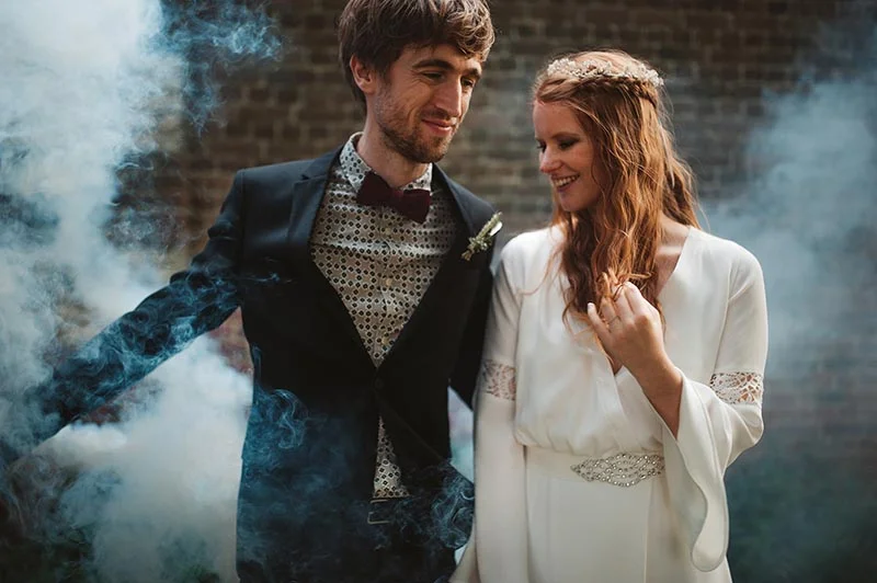 Jeunes mariés souriants dans une ambiance brumeuse, elle en robe blanche et lui en costume noir avec nœud papillon.