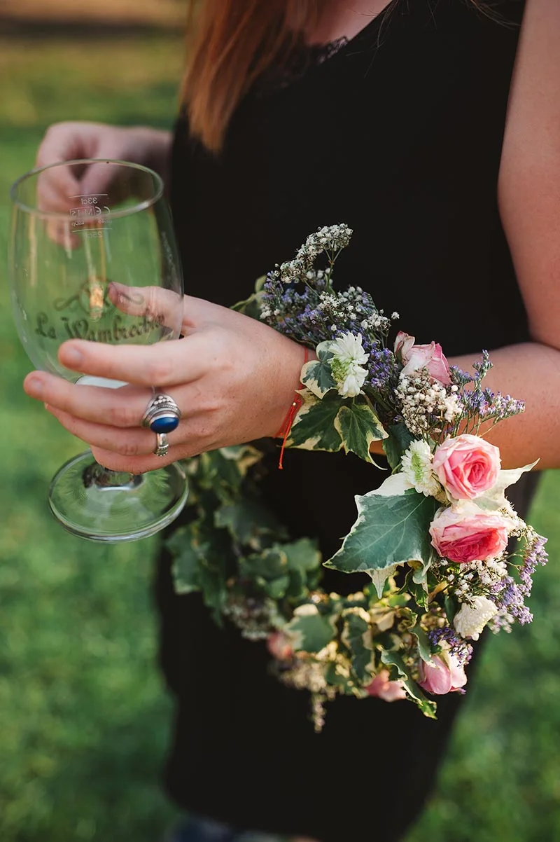 Une femme en robe noire tenant un verre et portant une couronne de fleurs champêtres et de lierre.