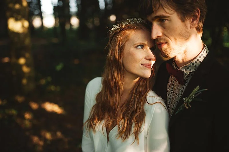 Couple de mariés complices dans une forêt baignée de lumière dorée, style bohème et naturel.