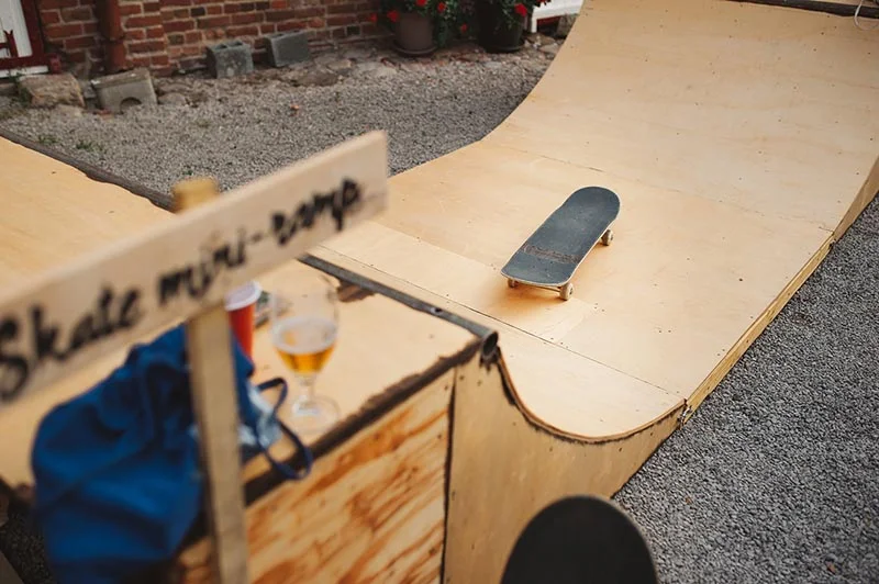 Un skateboard posé sur une mini-rampe artisanale en bois, avec un verre et un gobelet à côté.