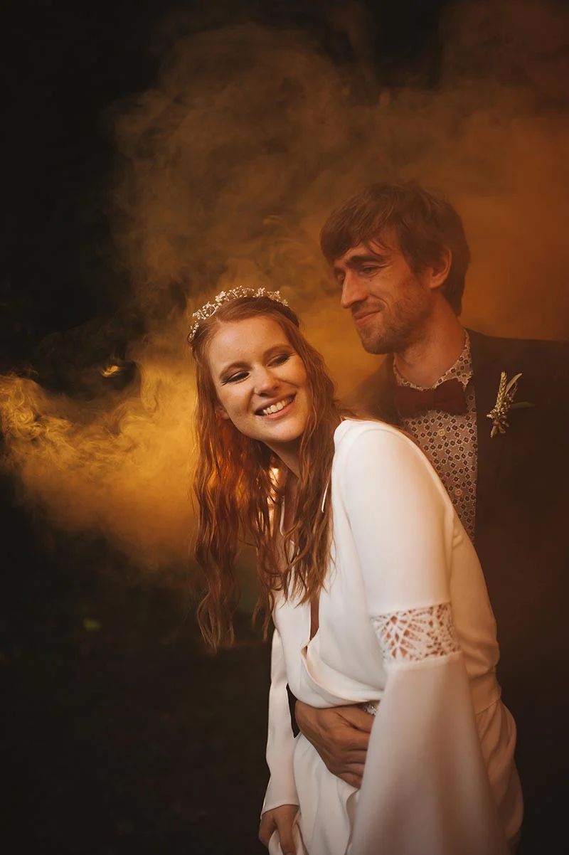 Couple souriant en tenue de mariage dans une ambiance brumeuse et chaleureuse aux tons dorés.