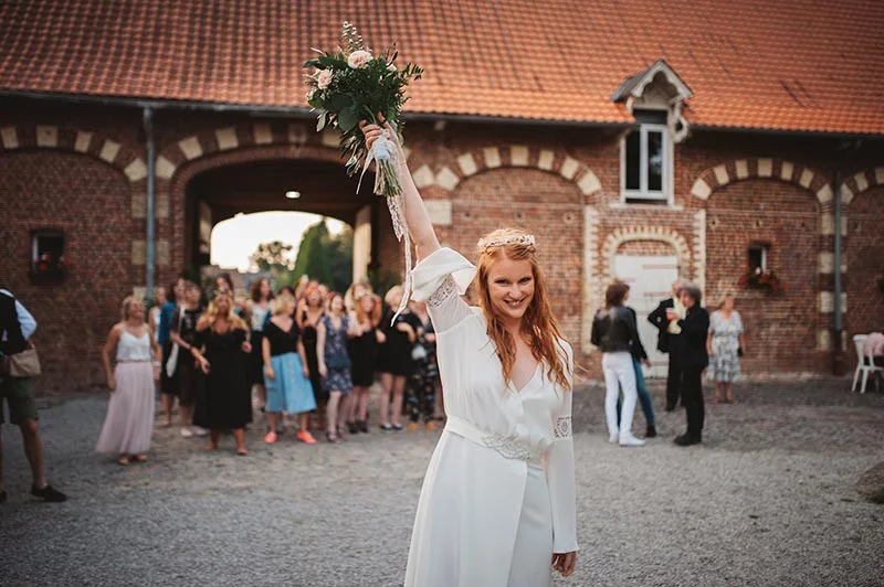Une mariée souriante en robe blanche brandit son bouquet devant ses invités et un bâtiment en briques rustiques.