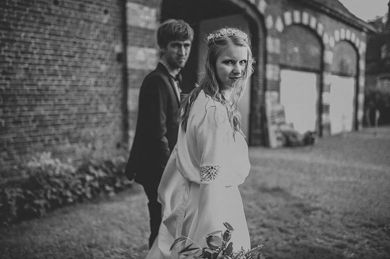 Couple de mariés en noir et blanc devant un bâtiment en briques, ambiance bohème et intimiste.