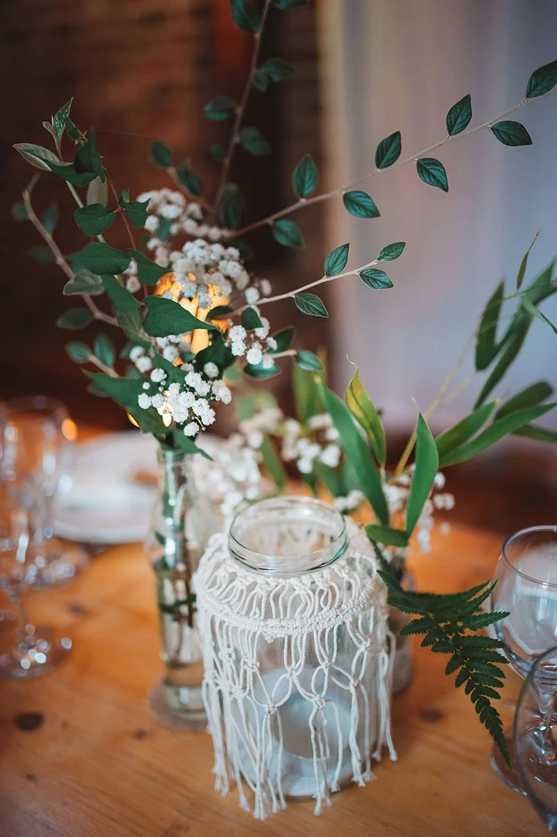 Décoration de table bohème avec vases en verre, fleurs blanches, feuillage vert et photophore en macramé sur bois clair.