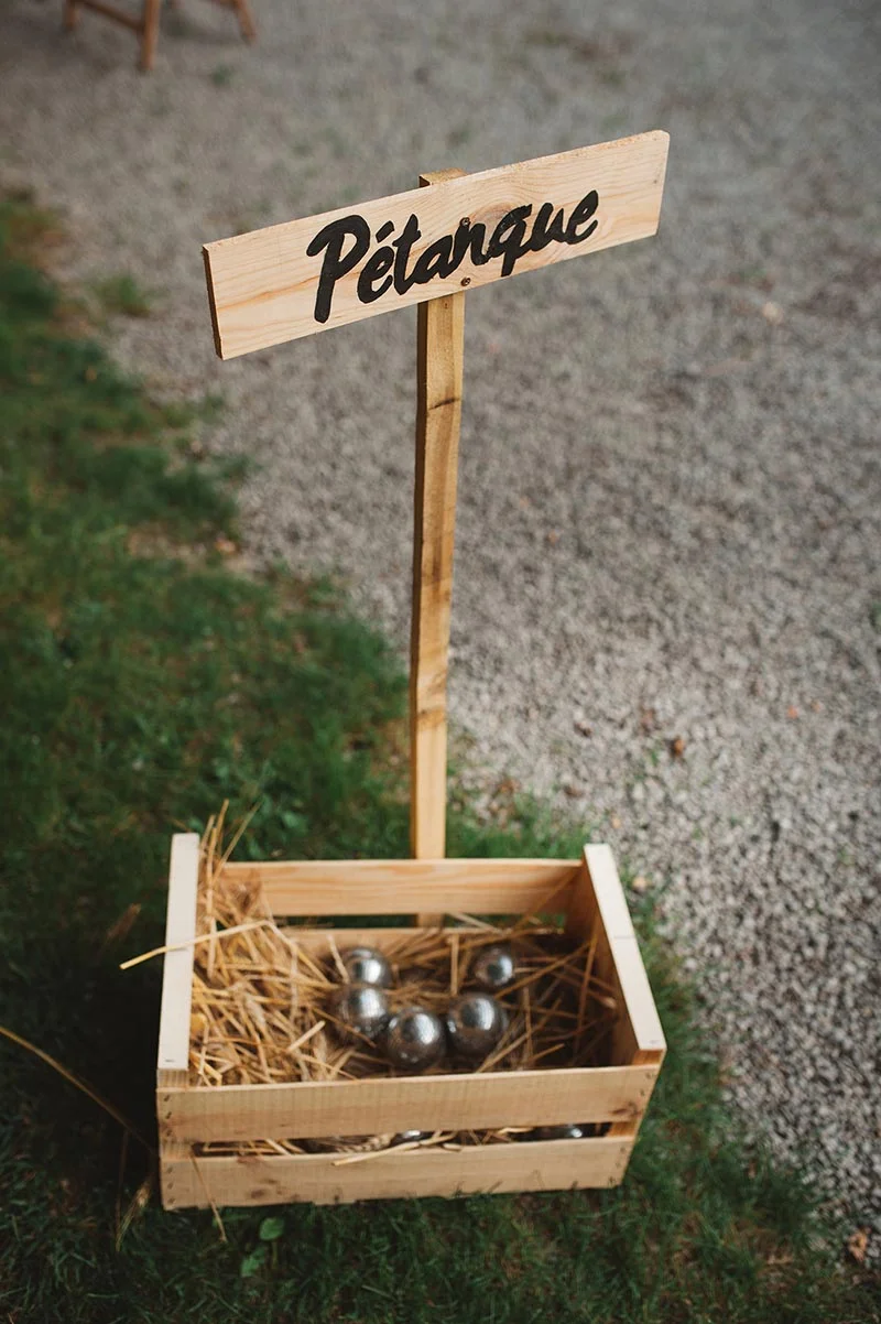 Caisse en bois contenant des boules de pétanque avec un panneau artisanal, posée sur l'herbe et le gravier.