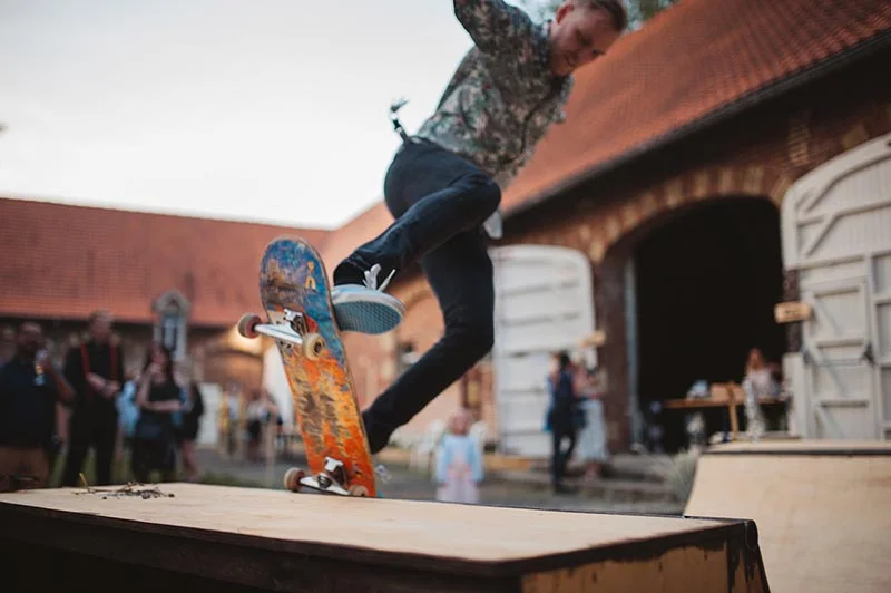 Un homme en tenue décontractée saute en skateboard devant des bâtiments en briques lors d'un mariage champêtre.