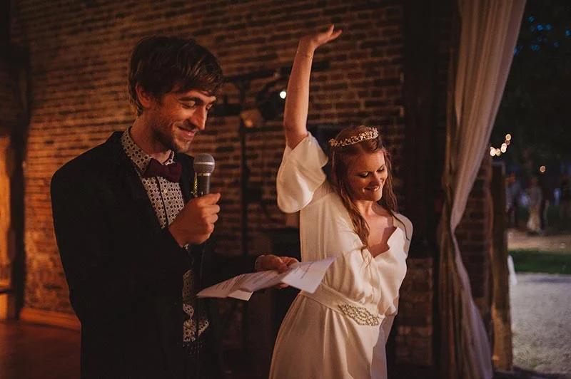 Un couple joyeux lors d'un discours, elle en robe blanche et lui en costume sombre, devant un mur de briques.