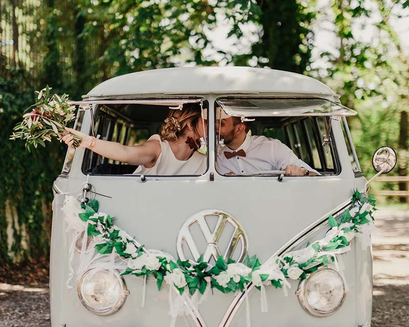 Jeunes mariés s'embrassant dans un Combi Volkswagen vintage blanc décoré de fleurs et de feuillage vert.