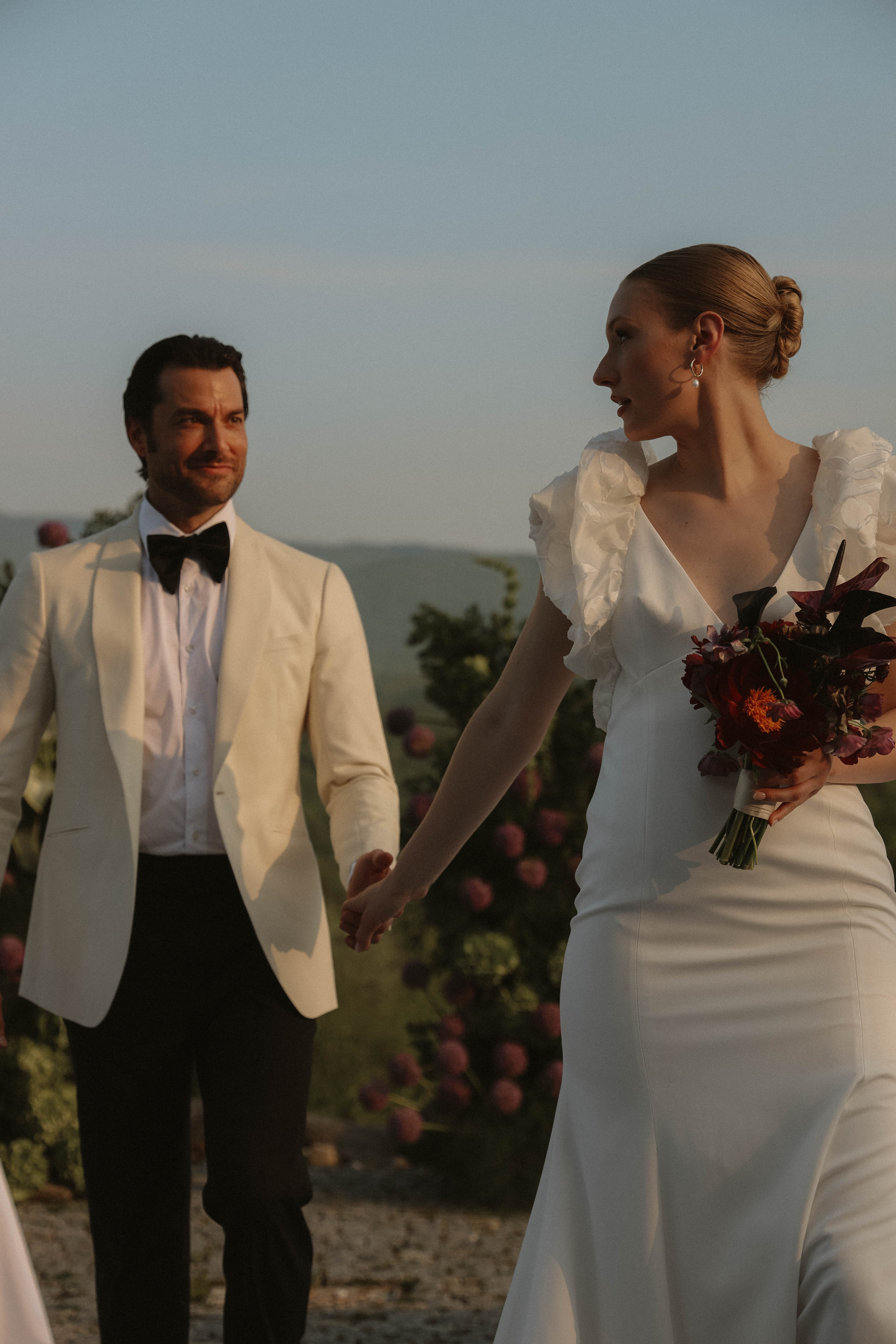Couple de mariés élégants main dans la main, robe blanche moderne et smoking crème, ambiance romantique au coucher du soleil.