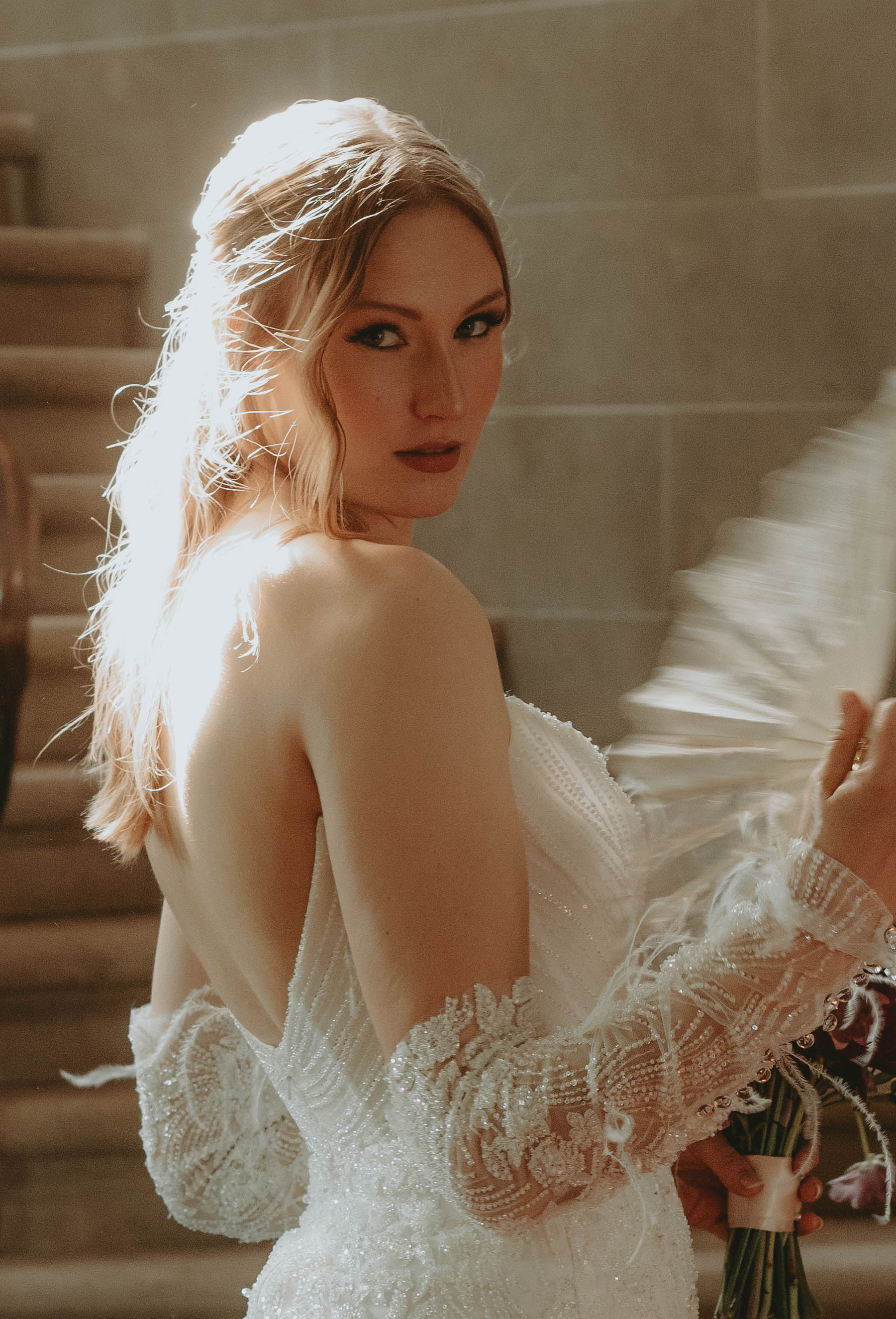 Jeune mariée blonde en robe blanche scintillante, regardant par-dessus son épaule dans une ambiance lumineuse et romantique.