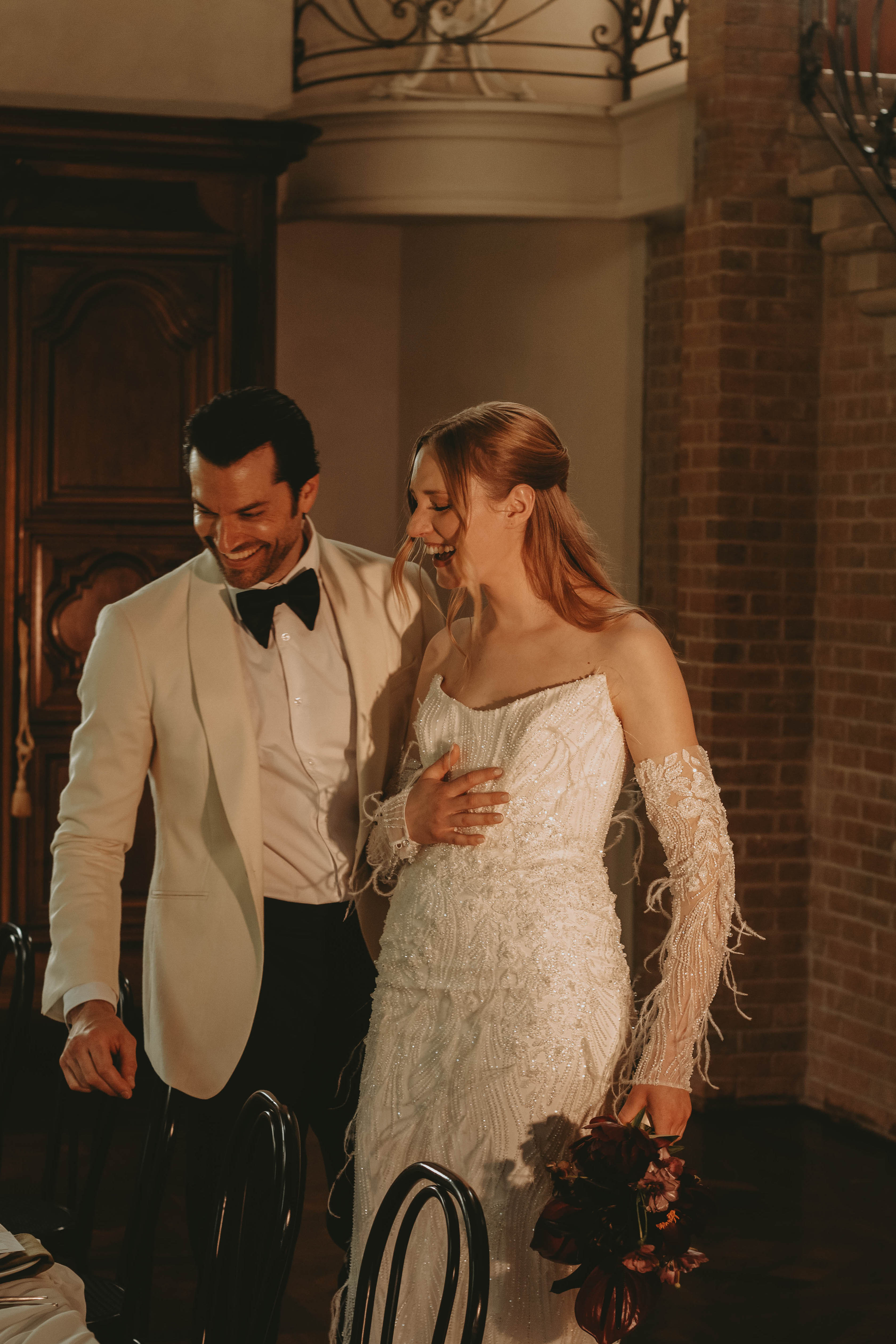 Couple riant, elle en robe blanche scintillante et lui en smoking blanc, dans un cadre chaleureux et romantique.