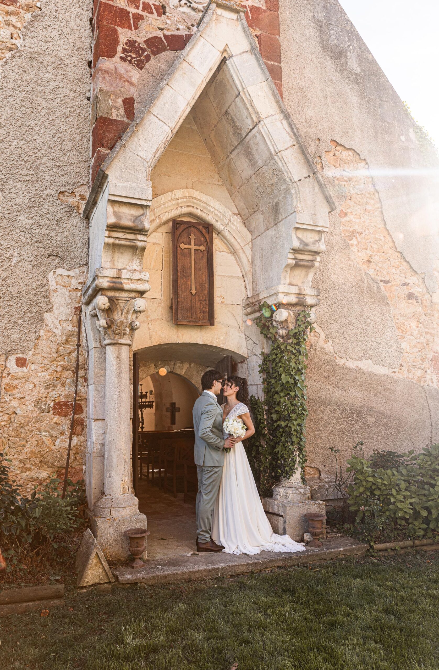 Jeunes mariés s'embrassant devant l'entrée en pierre d'une chapelle ancienne dans une ambiance romantique et lumineuse.