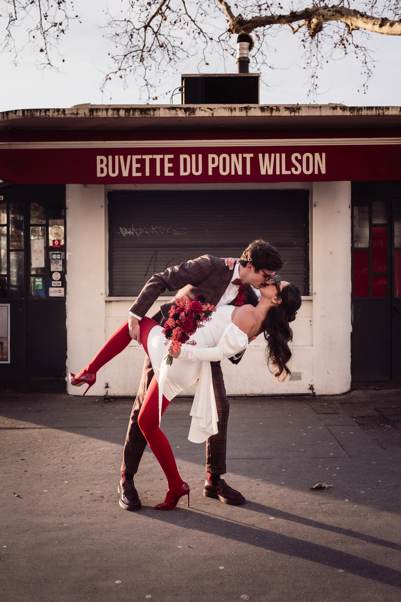Couple en tenue rétro chic s'embrassant devant la Buvette du Pont Wilson, avec des touches de rouge vif et une ambiance urbaine.