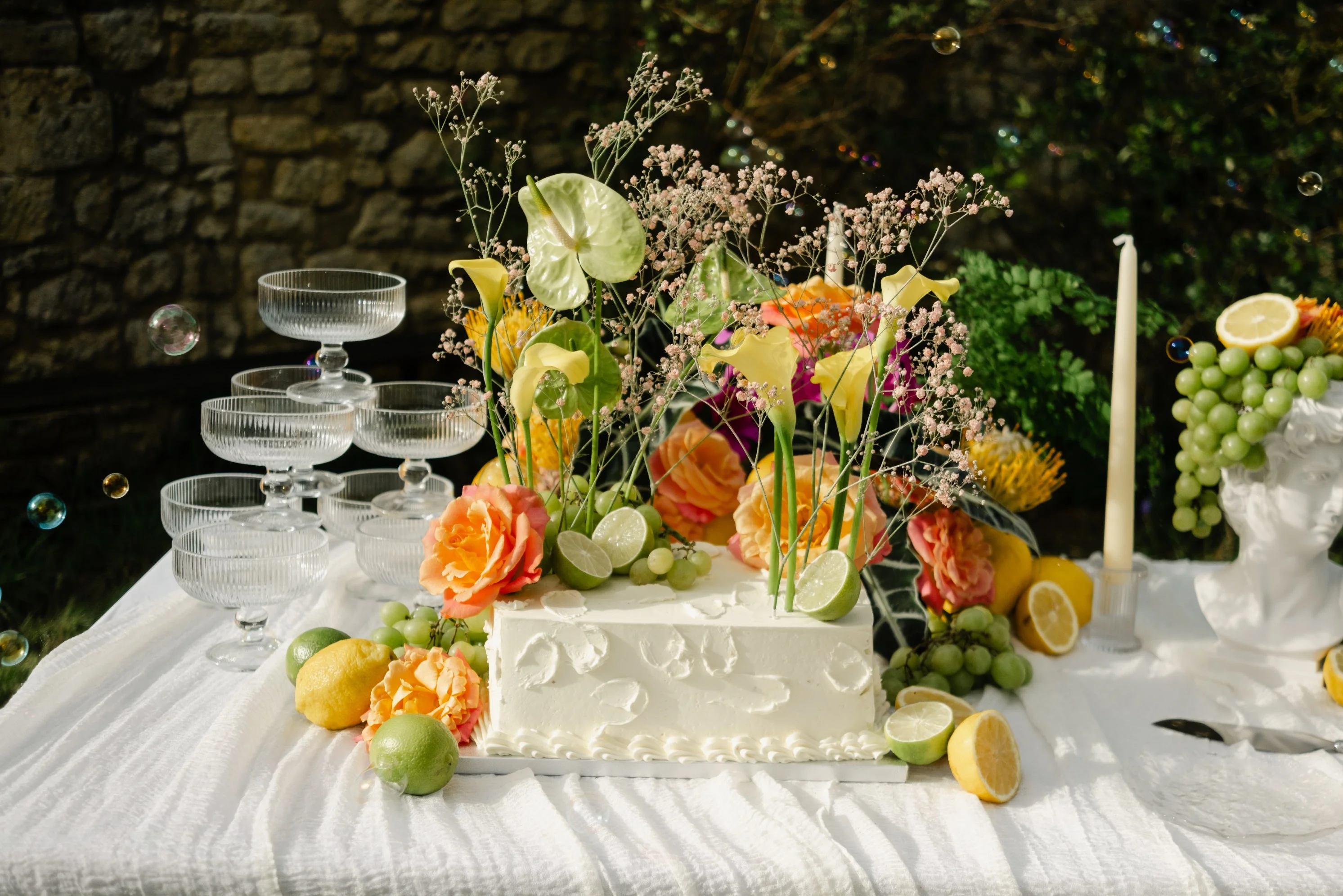 Table festive avec gâteau blanc, fleurs colorées, citrons et coupes en verre sur une nappe blanche en extérieur.