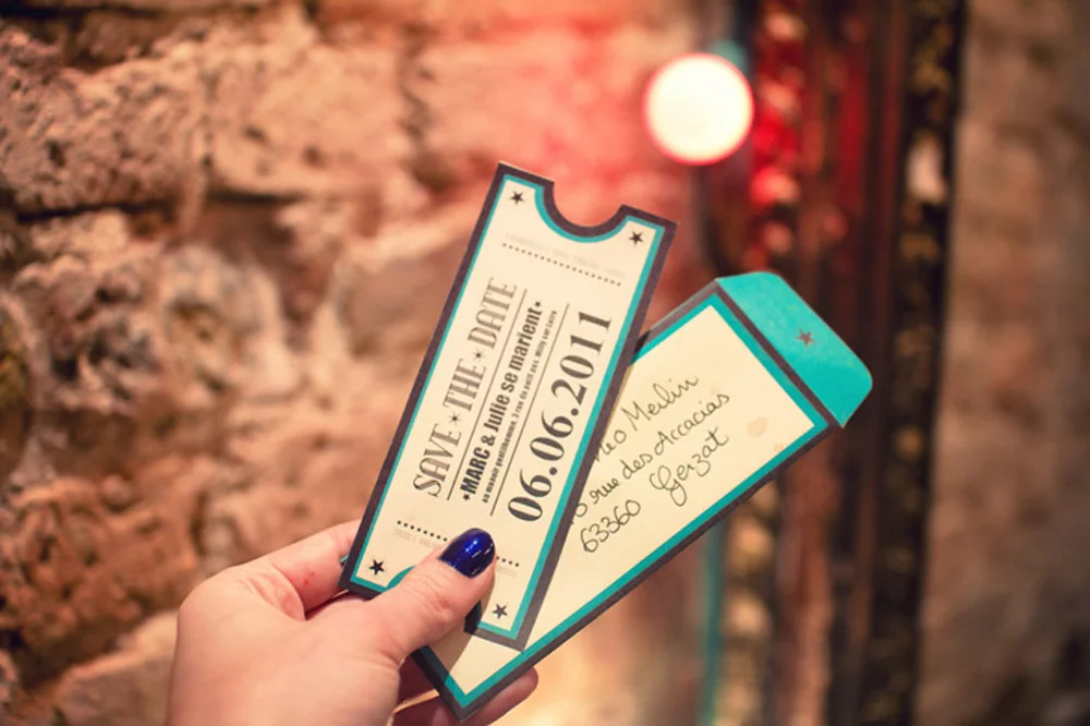 Main tenant un faire-part de mariage vintage en forme de ticket de concert bleu et blanc sur fond de mur en pierre.