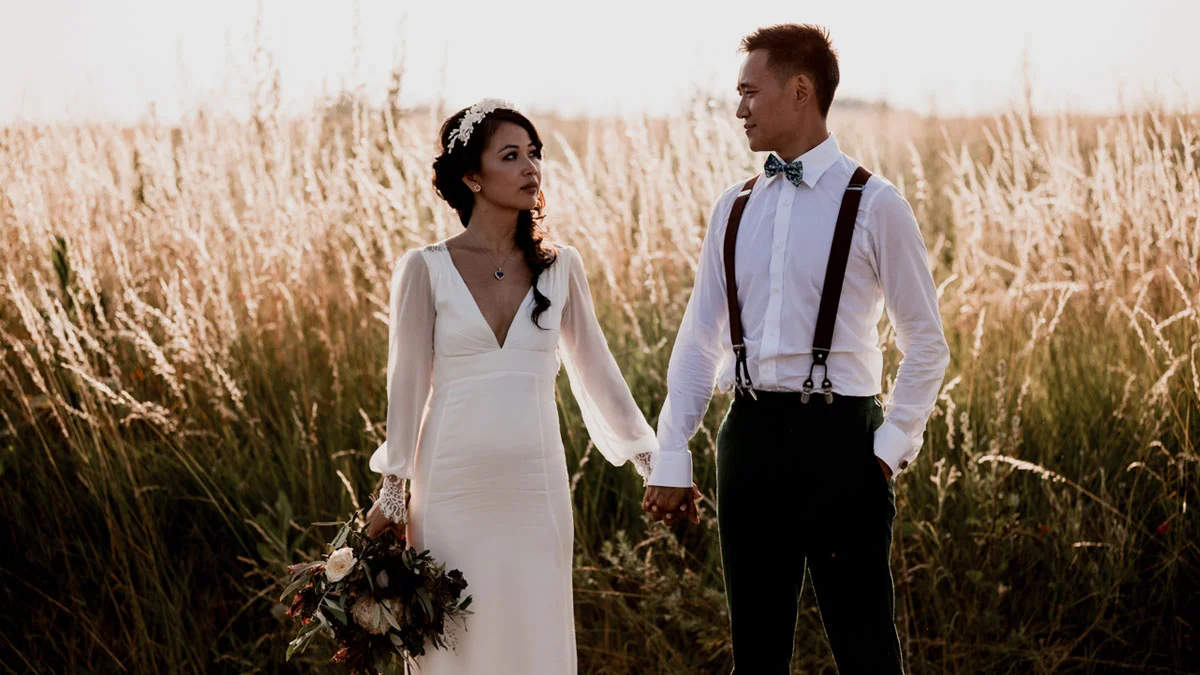 Couple en tenue de mariage bohème se tenant la main dans un champ d'herbes hautes au coucher du soleil.