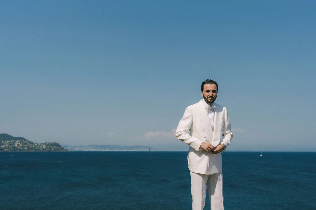 Un homme élégant en costume blanc et nœud papillon pose devant l'océan bleu sous un ciel clair.