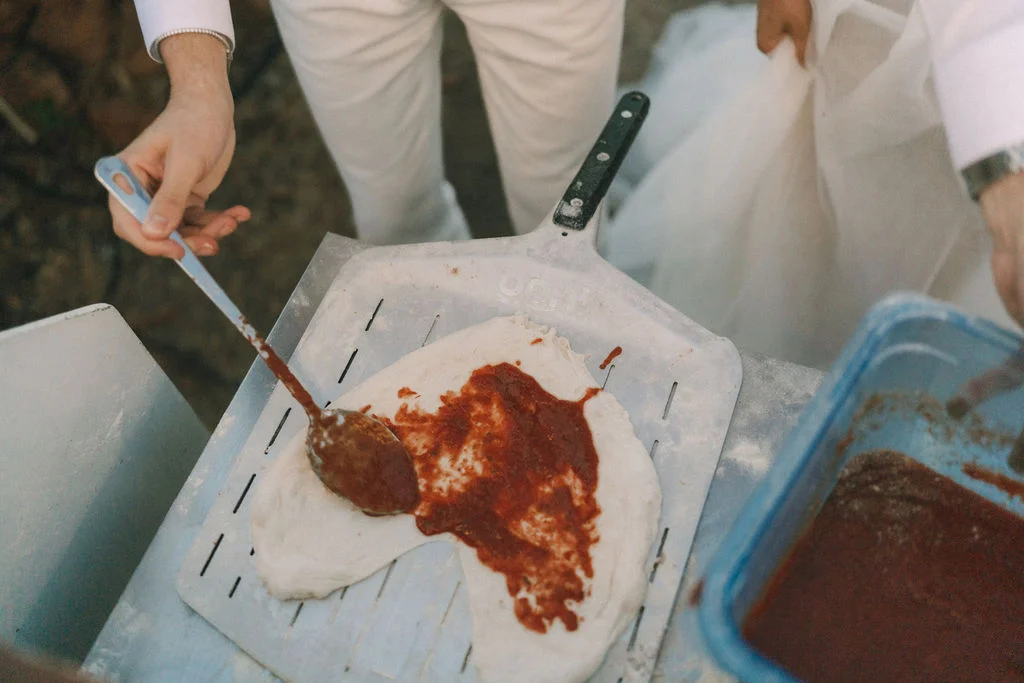 Un couple en tenue de mariage étale de la sauce tomate sur une pâte à pizza avec une cuillère.