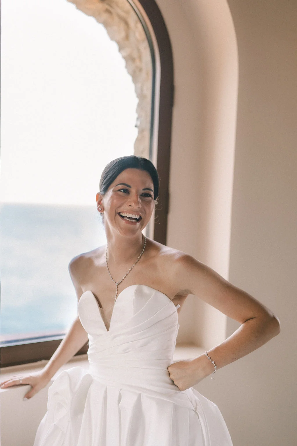 Une mariée souriante en robe blanche bustier et collier scintillant, posant près d'une fenêtre lumineuse.