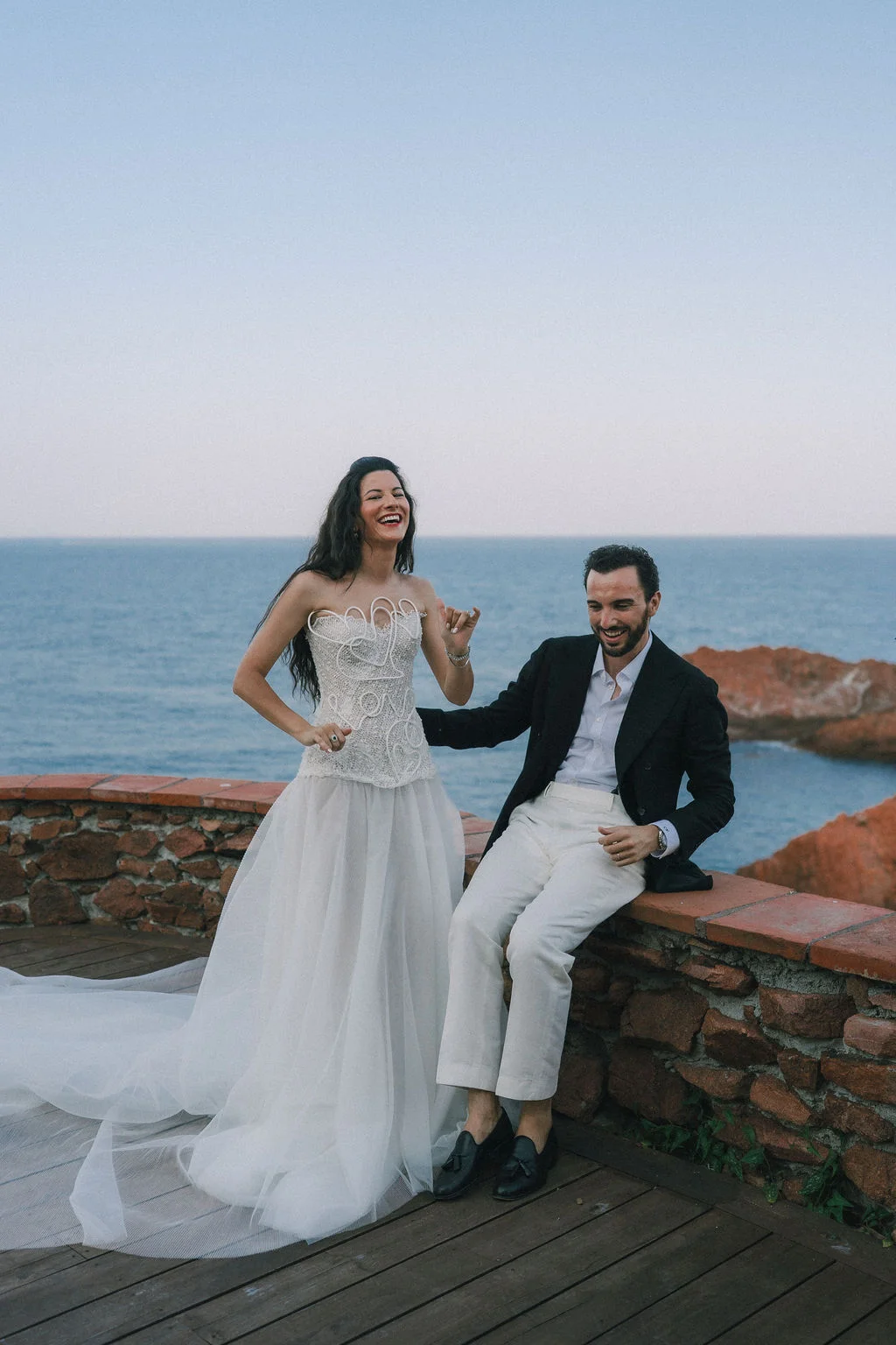 Jeunes mariés riant sur un muret face à la mer, elle en robe blanche stylée et lui en costume noir et pantalon blanc.