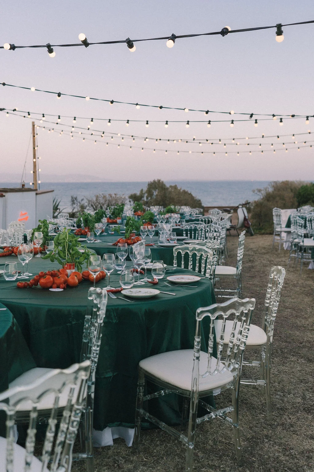 Table ronde nappée de vert avec tomates fraîches et chaises transparentes sous des guirlandes lumineuses face à la mer.