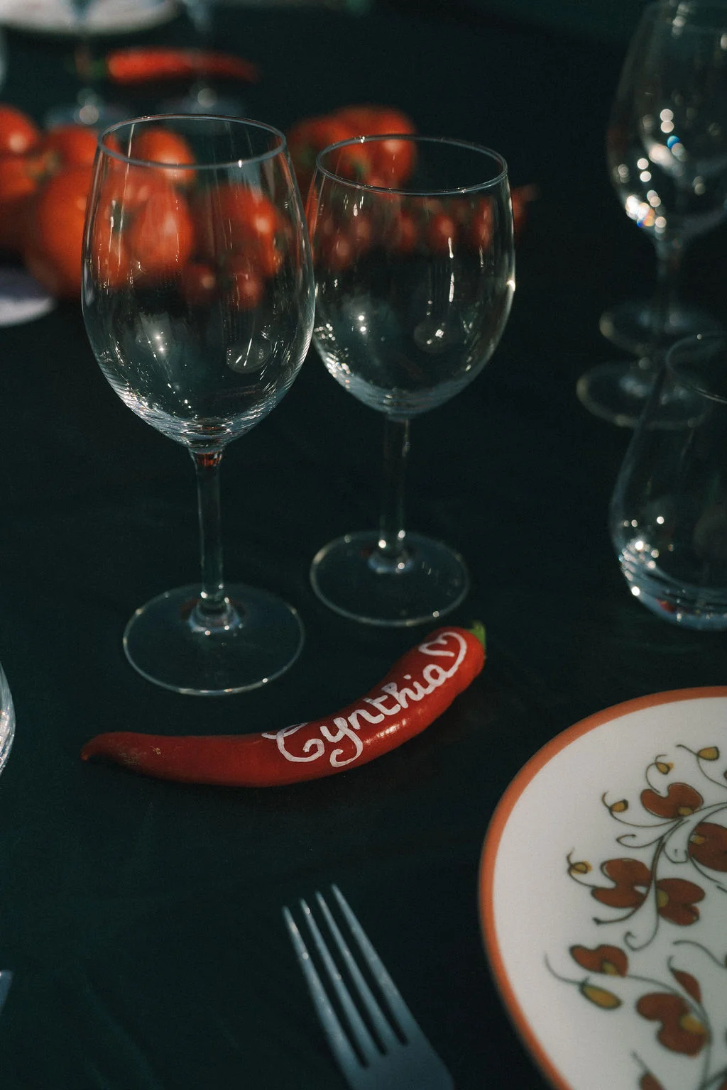 Détail d'une table sombre avec verres à pied et un piment rouge marqué du prénom Cynthia.