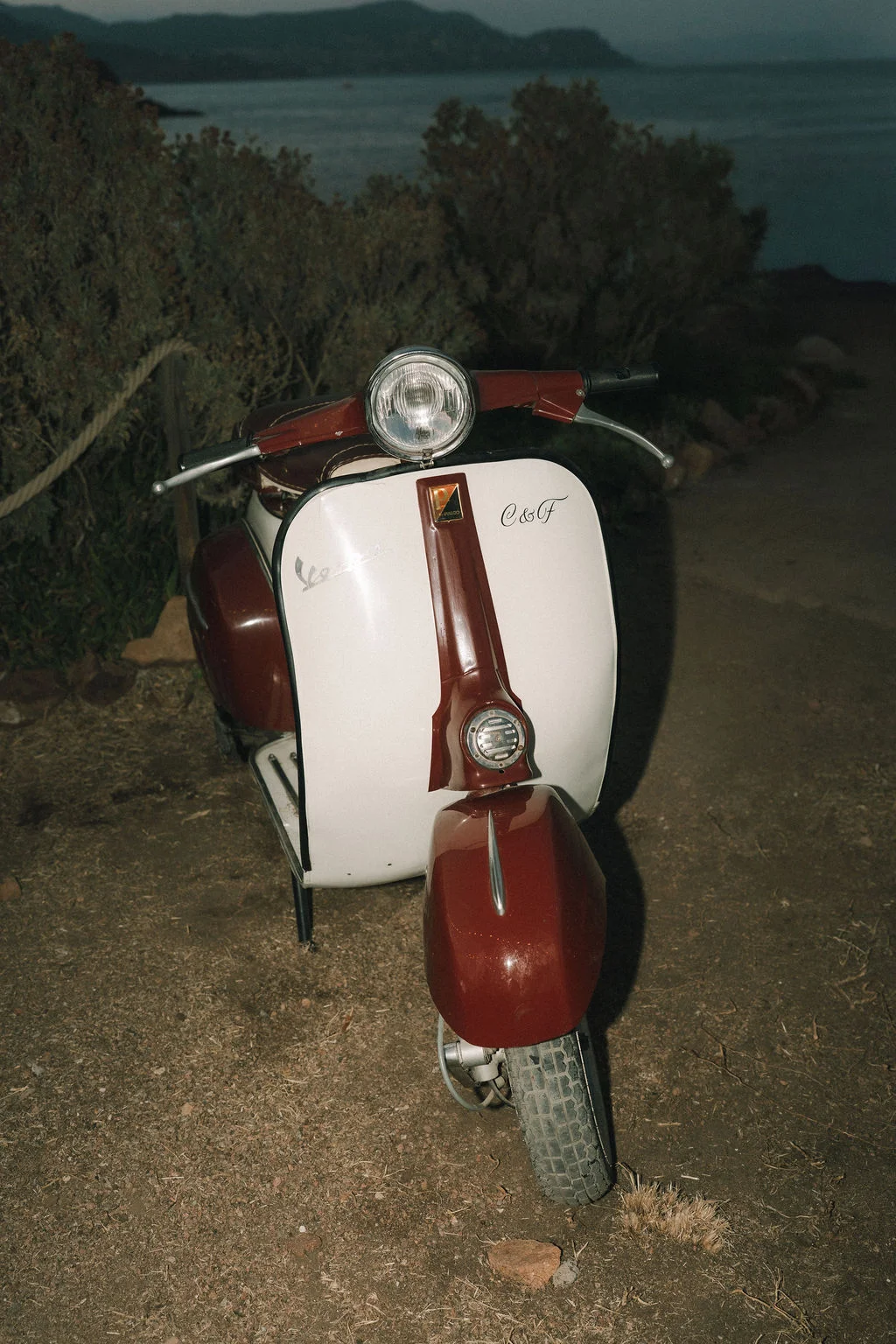 Scooter vintage blanc et bordeaux avec les initiales C&F, garé sur un chemin de terre face à la mer au crépuscule.