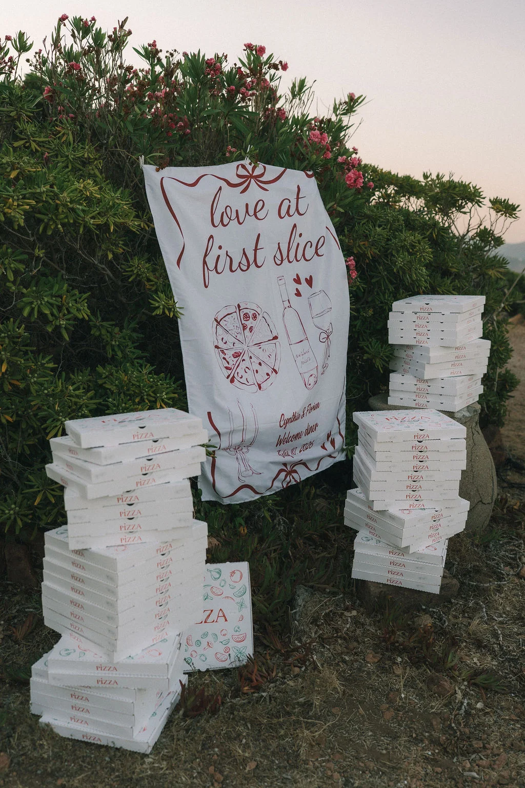 Piles de boîtes à pizza blanches devant un panneau "Love at first slice" dans un jardin luxuriant au coucher du soleil.