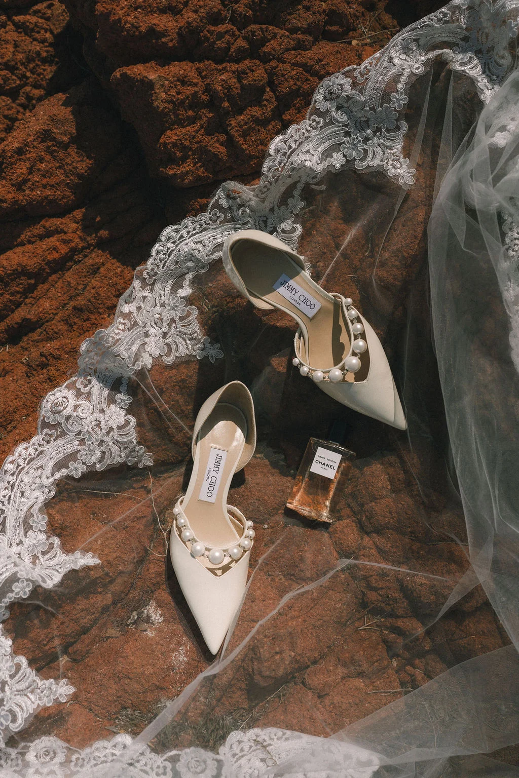Chaussures Jimmy Choo blanches et parfum Chanel posés sur un voile en dentelle et des rochers rouges.