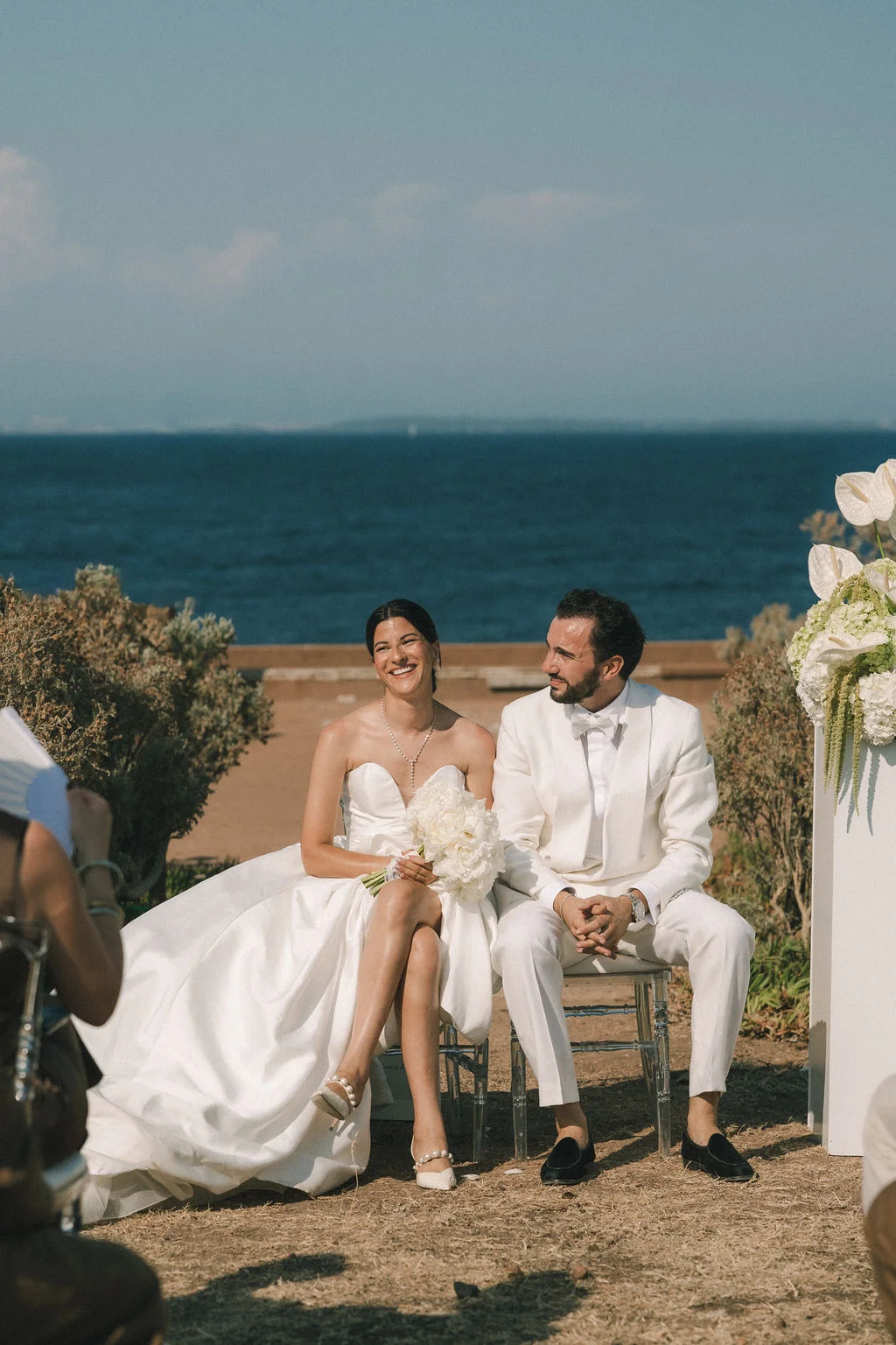 Jeunes mariés élégants en blanc assis face à la mer, ambiance lumineuse et romantique sur la côte.