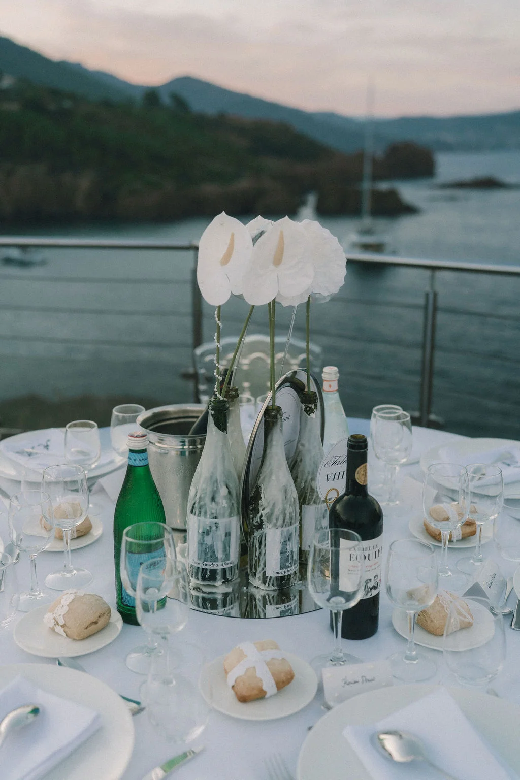 Table élégante avec fleurs blanches et vin, dressée face à une vue panoramique sur la mer au crépuscule.