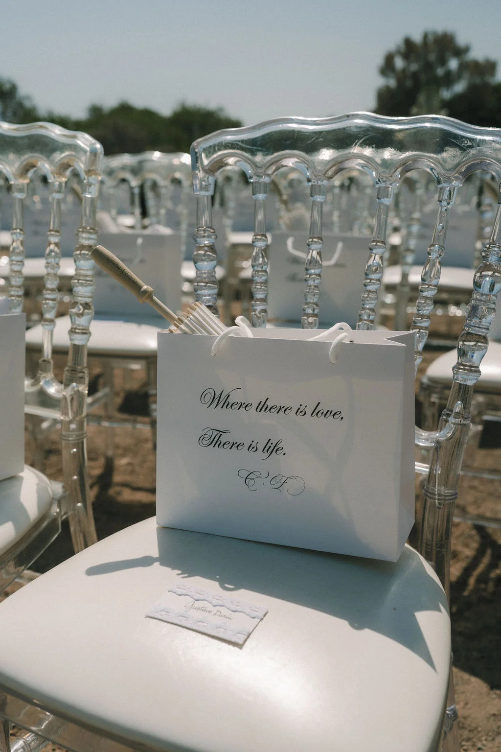 Sac cadeau blanc avec citation romantique posé sur une chaise transparente lors d'un mariage élégant en extérieur.