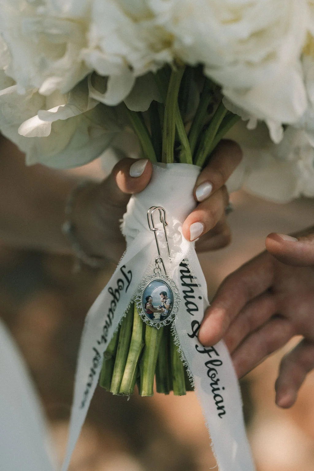 Gros plan sur un bouquet de roses blanches tenu par les mariés, orné d'un médaillon photo et d'un ruban personnalisé.