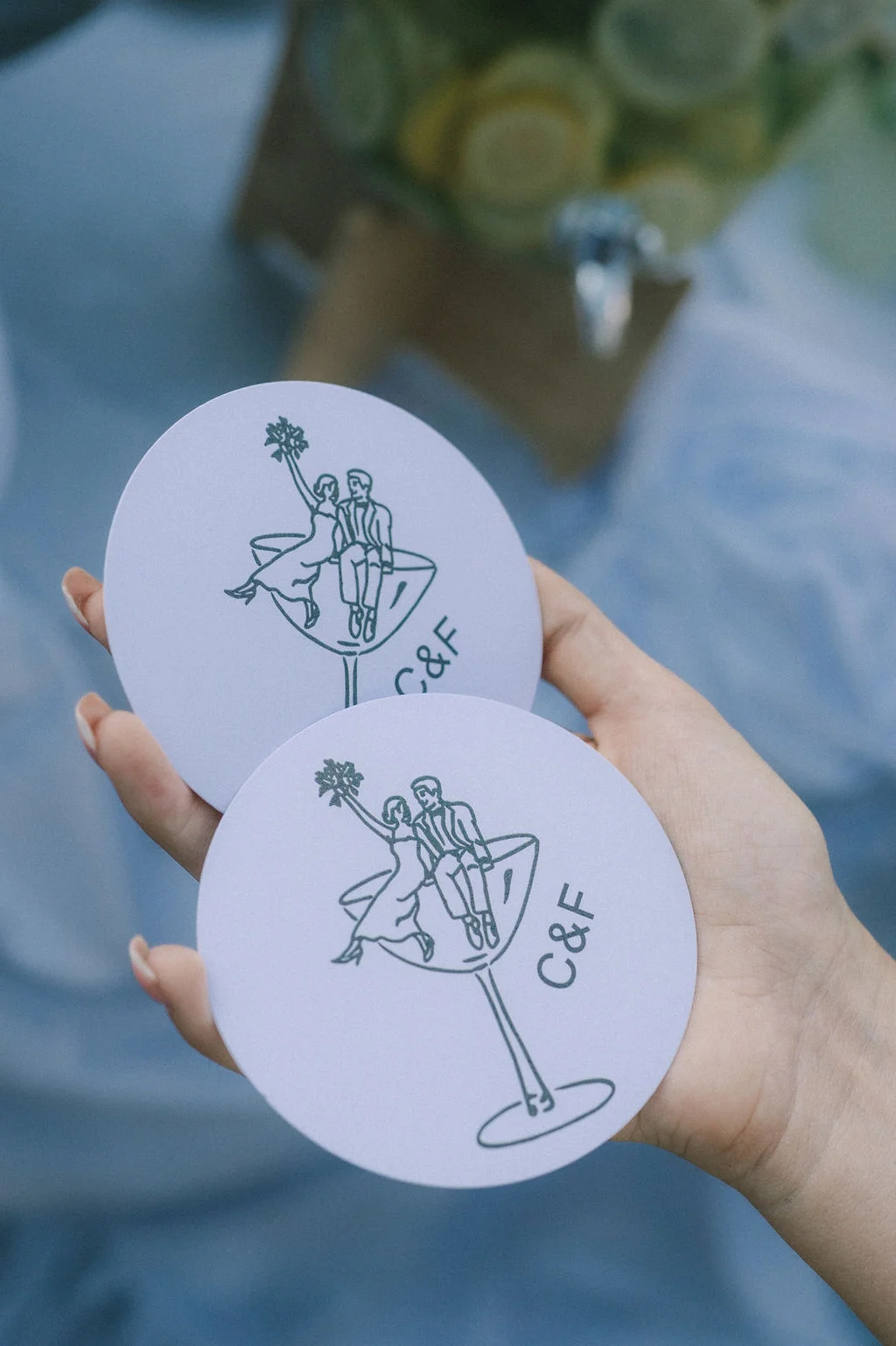 Main tenant deux sous-verres blancs circulaires avec une illustration minimaliste de couple et les initiales C&F.