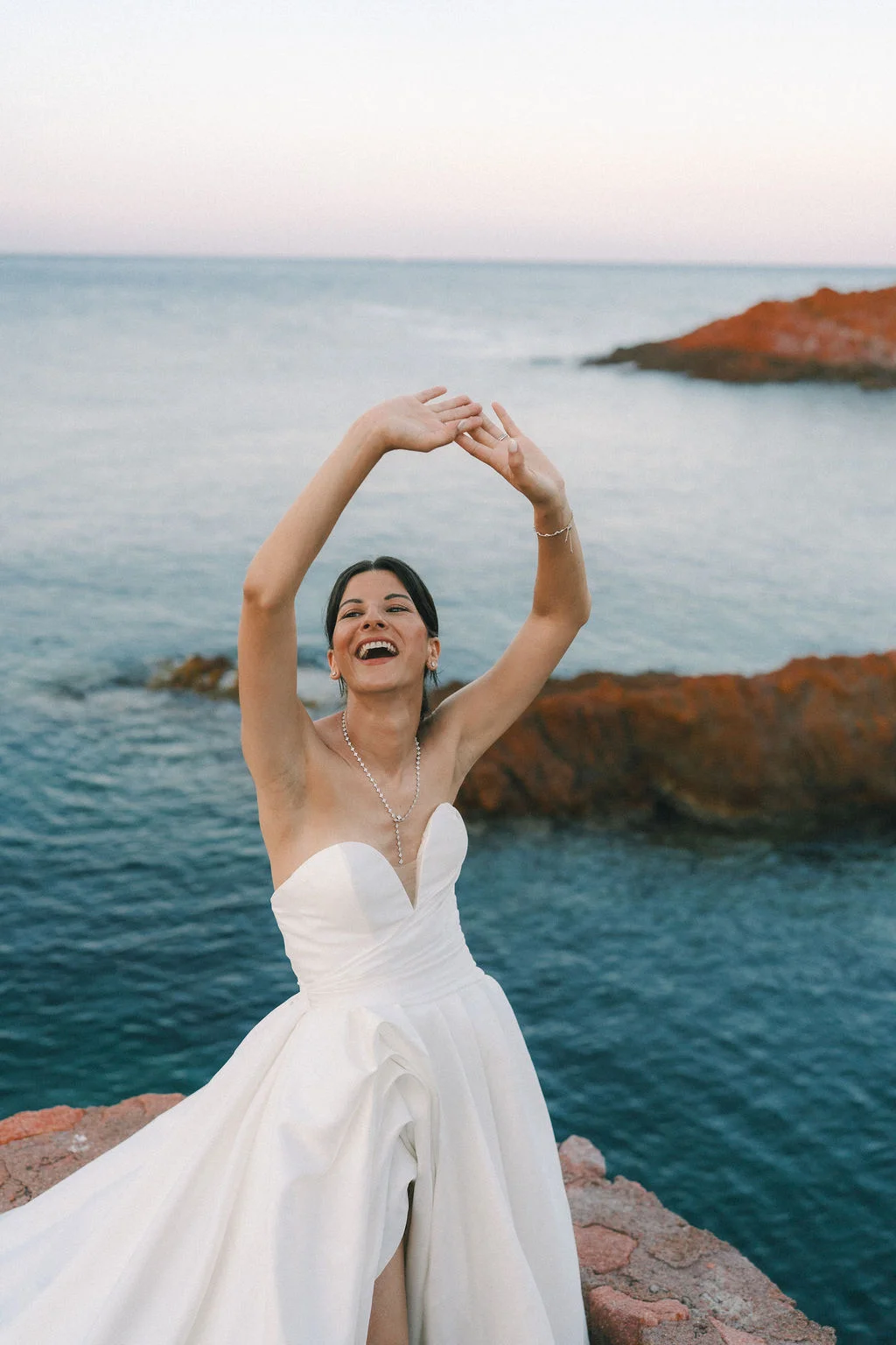 Une mariée rayonnante en robe blanche rit les bras levés face à la mer sur les rochers du Cap Roux.