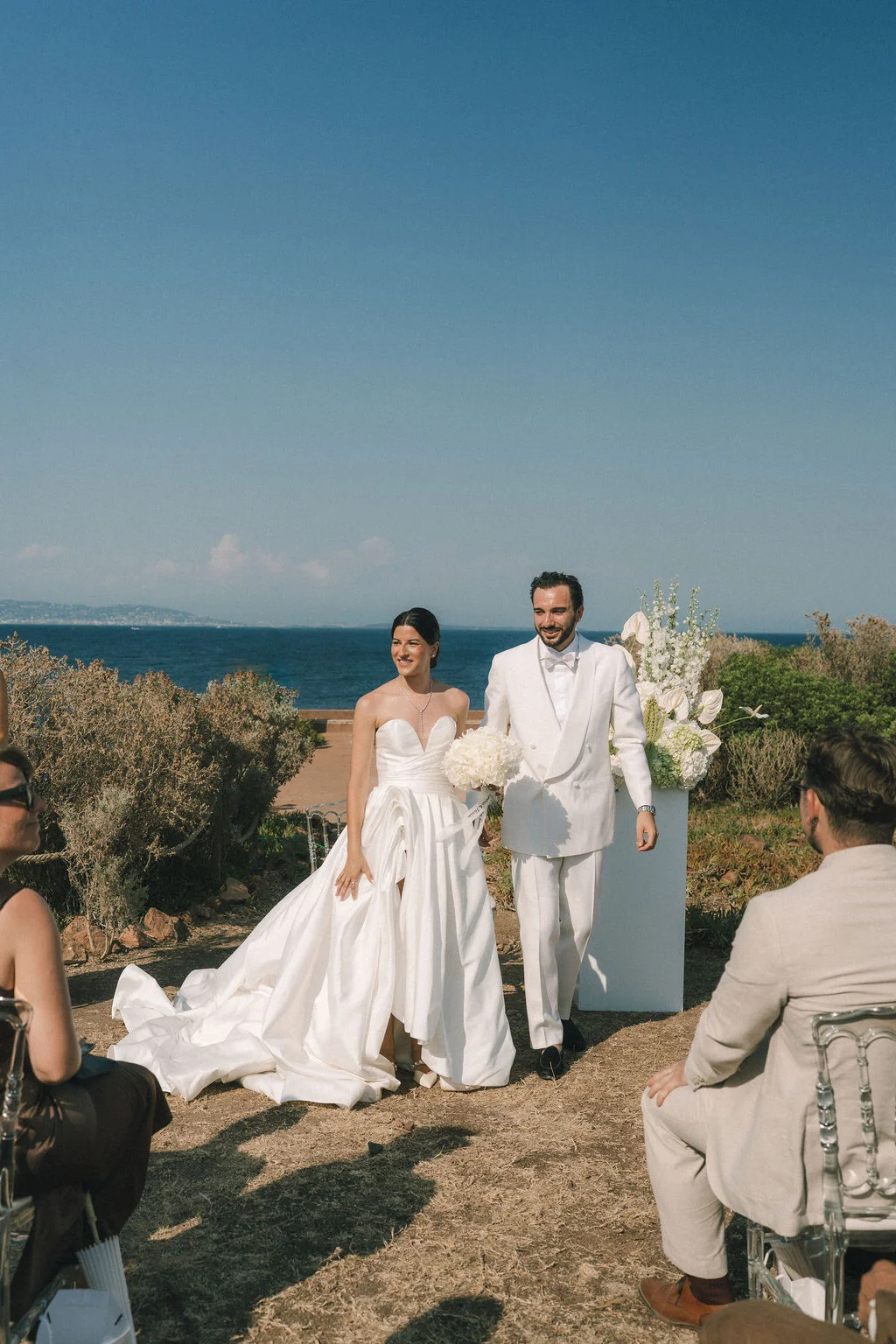Jeunes mariés élégants en blanc posant en extérieur face à la mer sous un ciel bleu lumineux.
