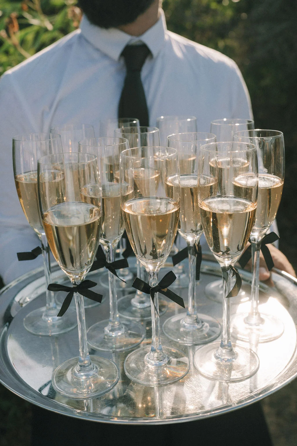 Un serveur en chemise blanche présente un plateau de coupes de champagne ornées de nœuds noirs pour un mariage élégant.