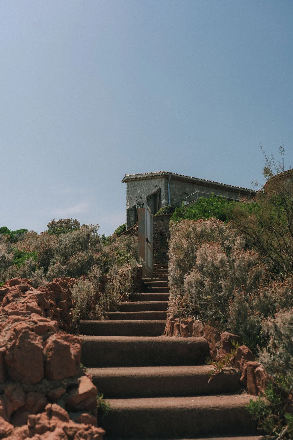 Escalier en pierre montant vers une bâtisse rustique entourée de végétation sauvage sous un ciel bleu clair.