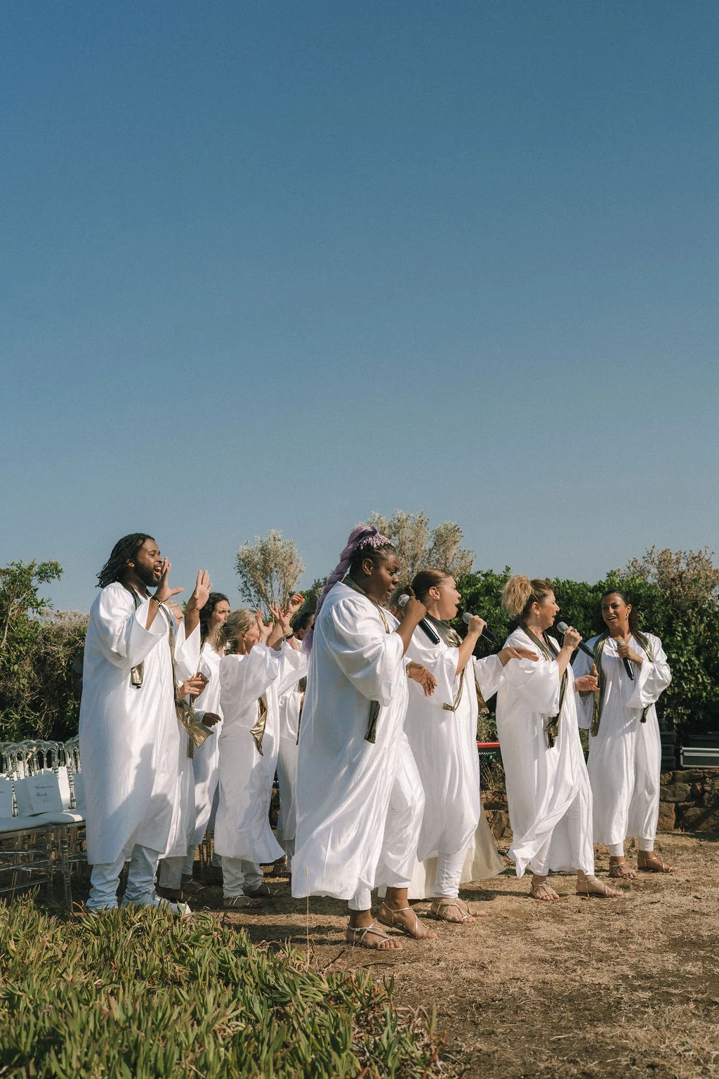 Un groupe de chanteuses en robes blanches et dorées performant en extérieur sous un ciel bleu dégagé.