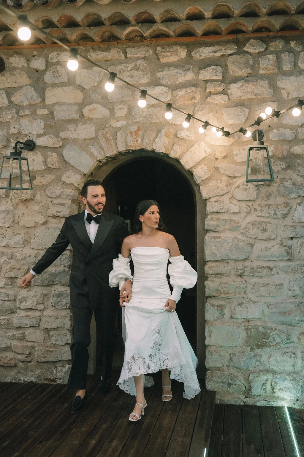Couple élégant sortant d'une arche en pierre sous des guirlandes lumineuses, ambiance romantique et chic.