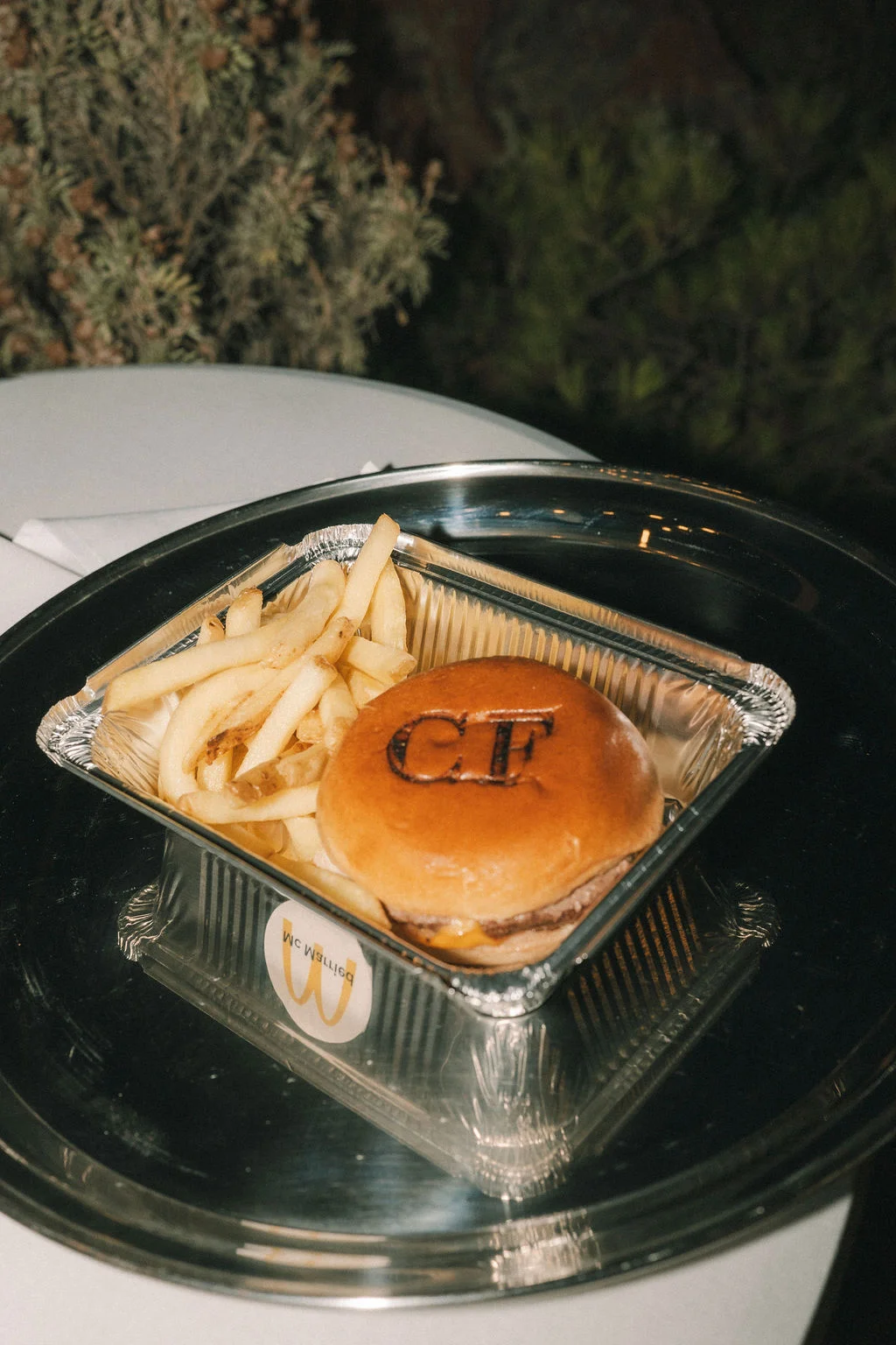 Un mini burger marqué CF et des frites dans un plat en aluminium posé sur un plateau argenté.