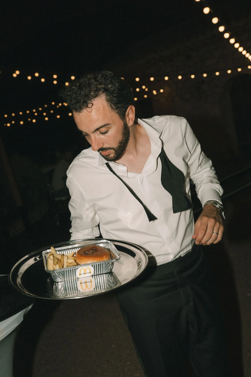Un serveur en chemise blanche apporte un burger et des frites sur un plateau lors d'une soirée de mariage nocturne.