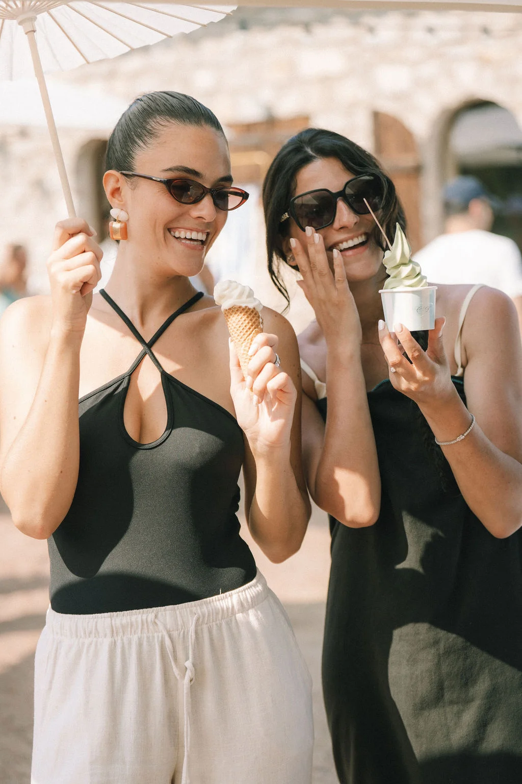 Deux femmes souriantes et stylées profitant d'une glace sous un parasol blanc dans une ambiance estivale et lumineuse.