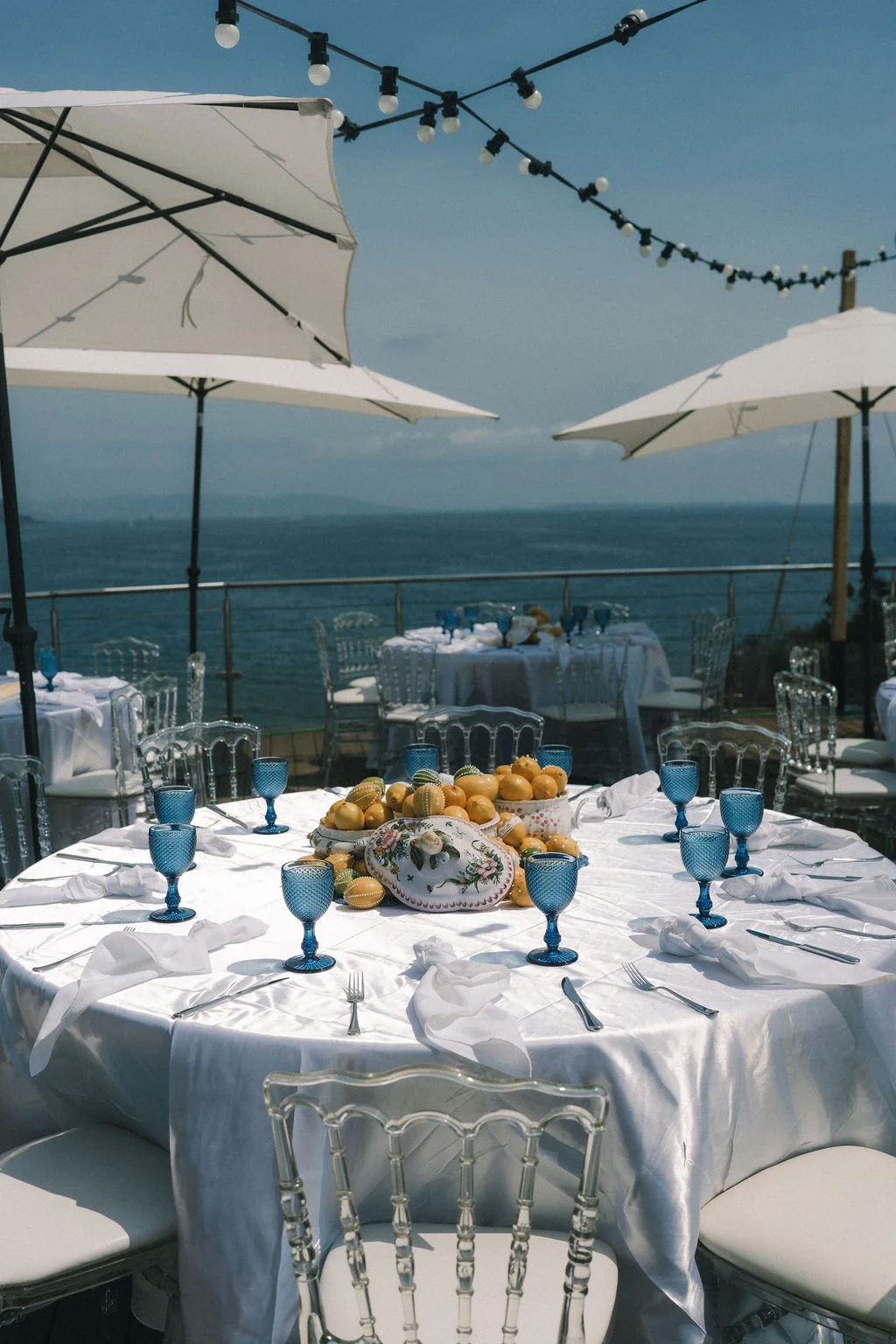 Table ronde élégante avec citrons et verres bleus sur une terrasse face à la mer sous des parasols blancs.