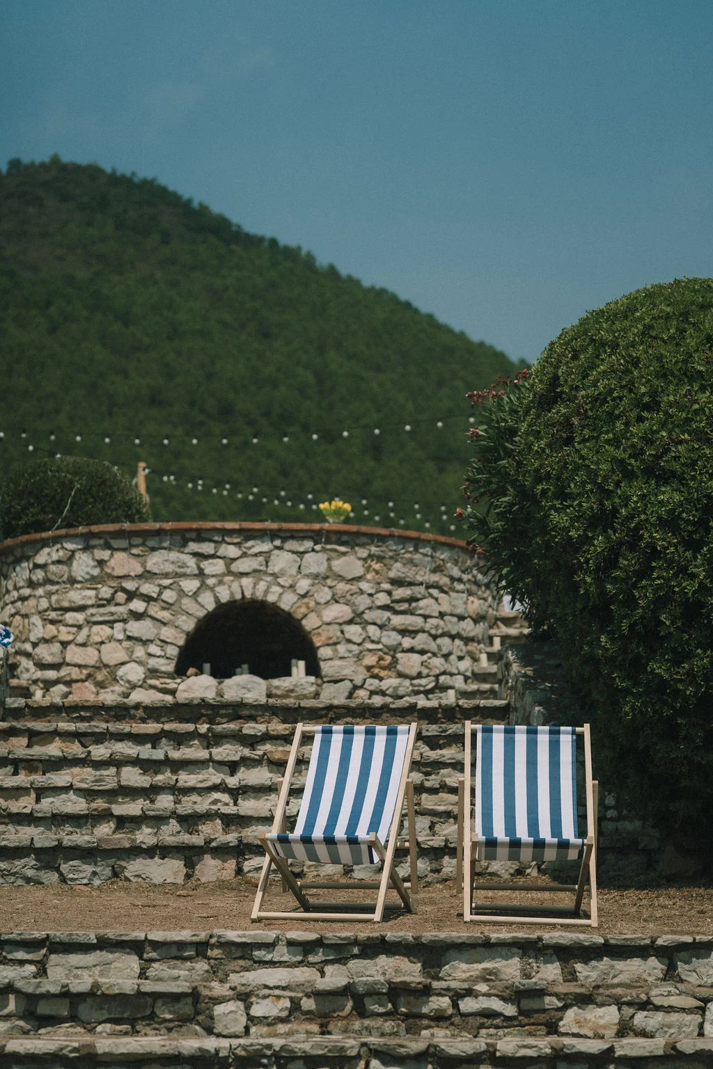 Deux chaises longues rayées bleu et blanc devant un mur de pierres et une colline verdoyante sous un ciel bleu.