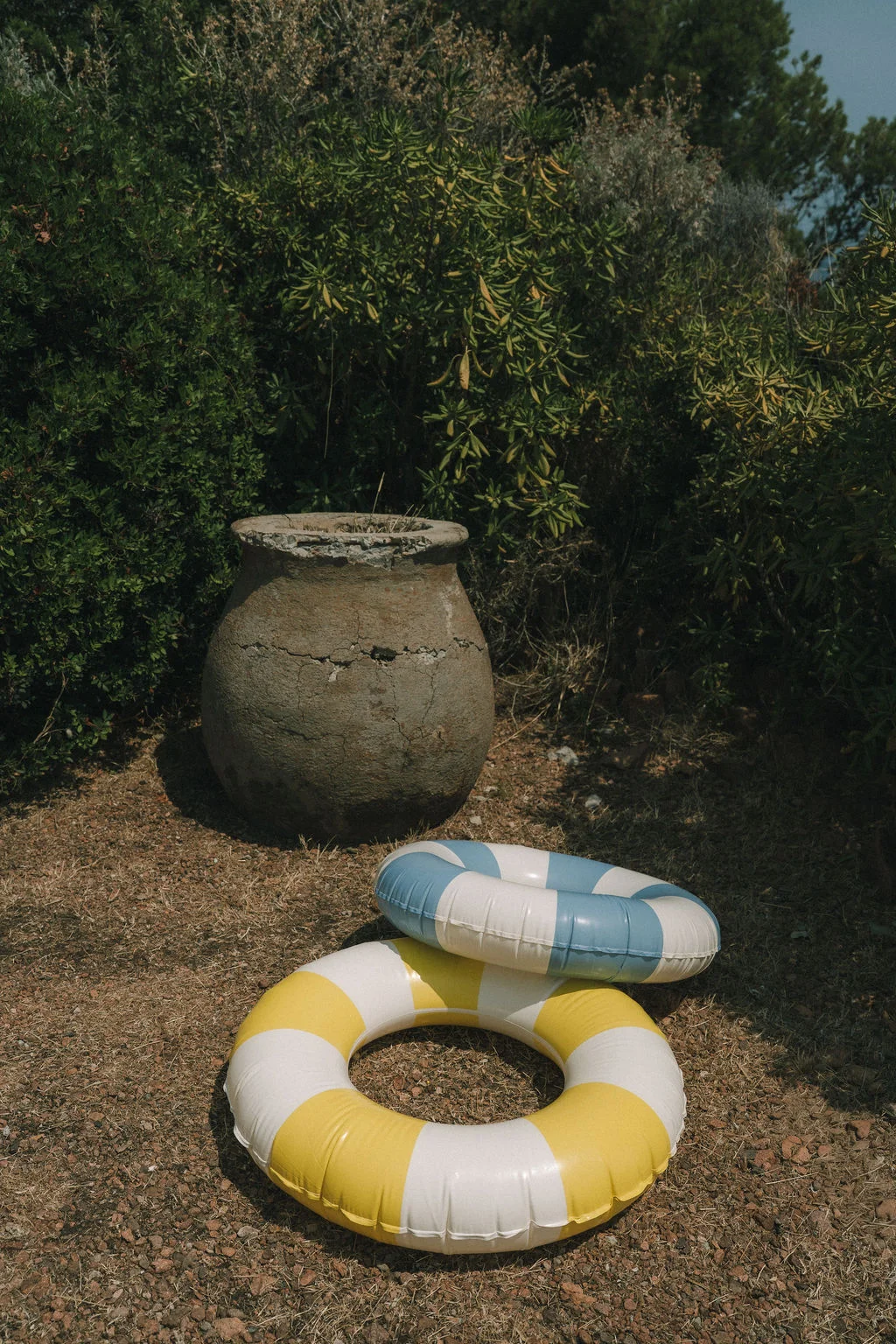 Deux bouées gonflables rayées jaune et blanc, et bleue et blanc, posées au sol devant une jarre ancienne et des buissons.