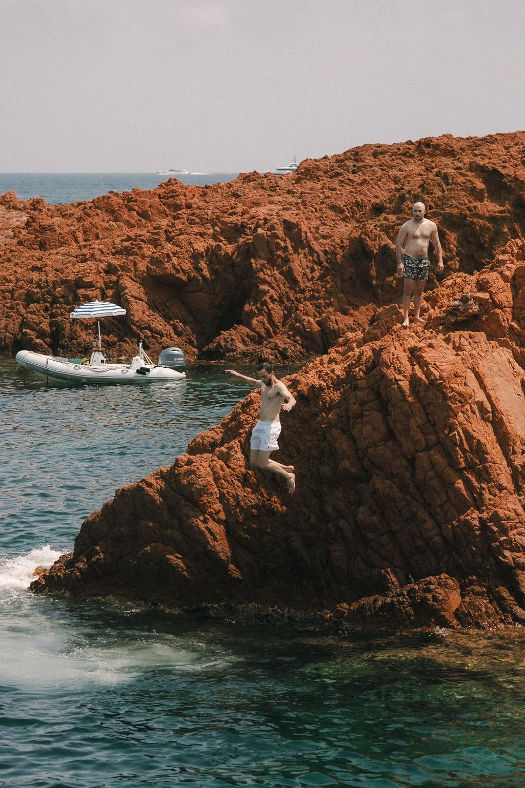 Un homme saute d'un rocher rouge dans une eau turquoise, tandis qu'un autre observe près d'un bateau pneumatique.